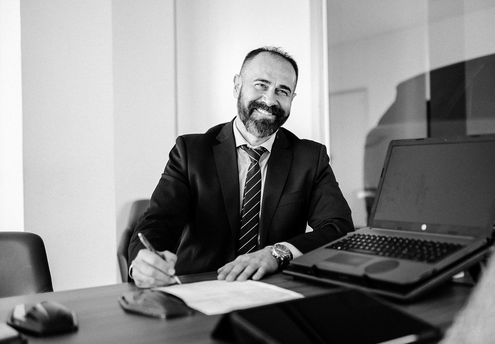 Man in suit and tie, sitting at desk with papers and laptop, smiling