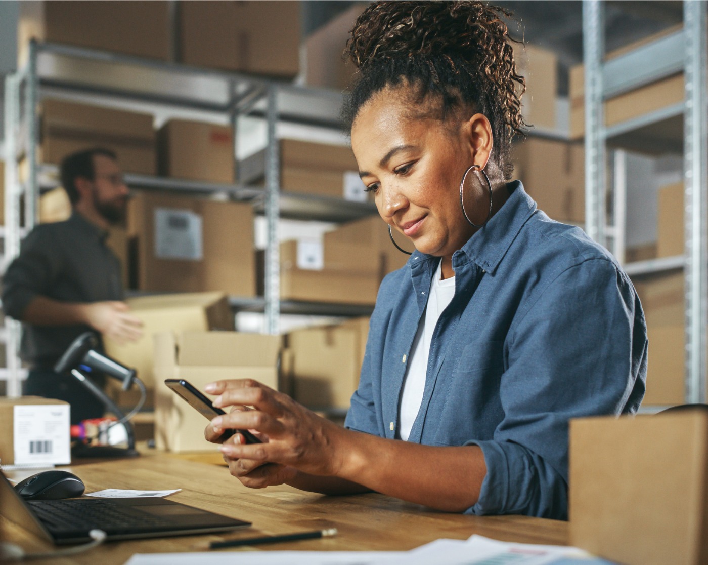 Gestionnaire d’inventaire utilisant un smartphone pour scanner un code-barres sur un colis, préparer une petite boîte en carton pour l’affranchissement. Propriétaire d’une petite entreprise multiethnique noire travaillant sur un ordinateur portable.