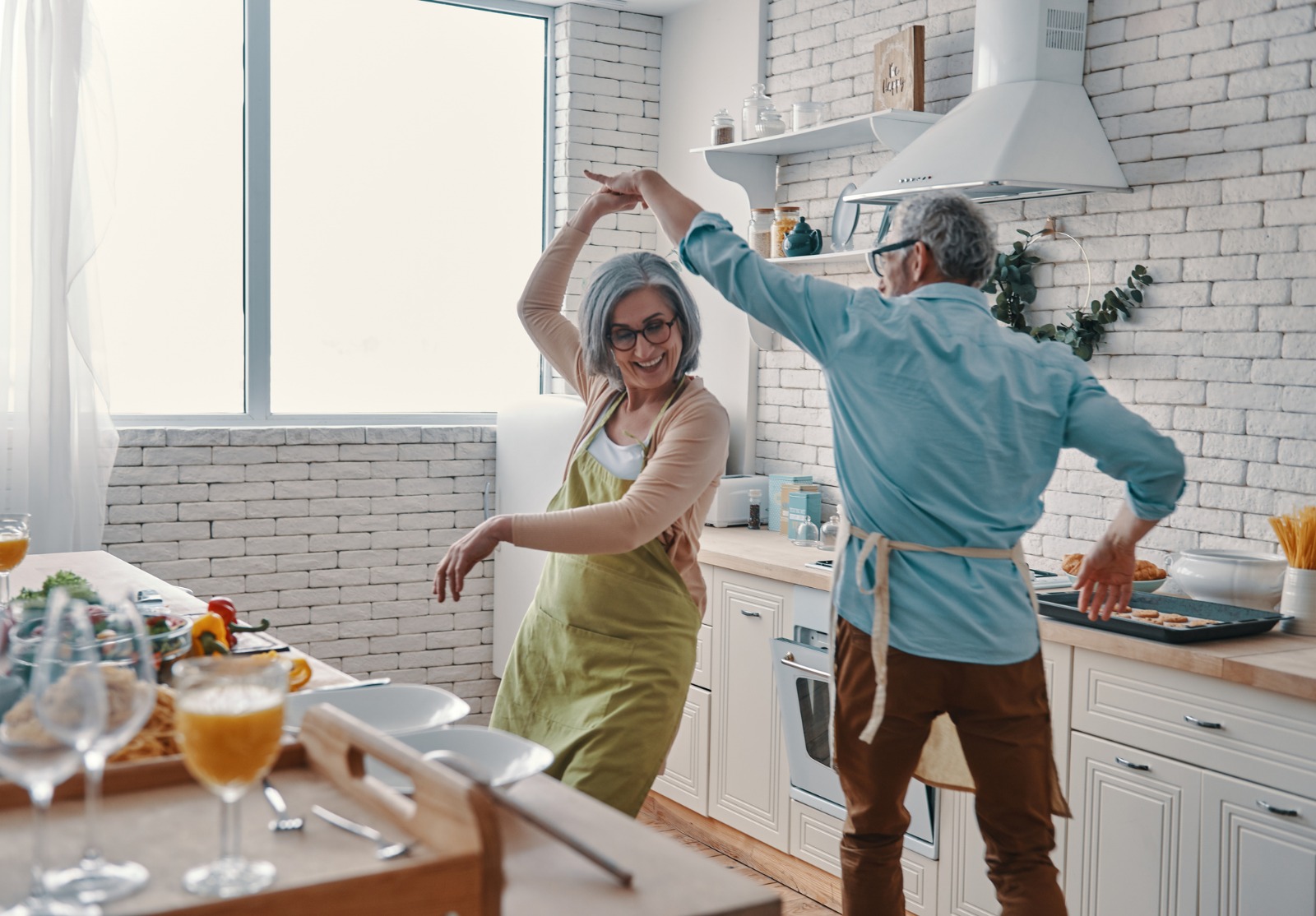 Beau couple d'aînés espiègles vêtus de tabliers dansant dans leur cuisine.
