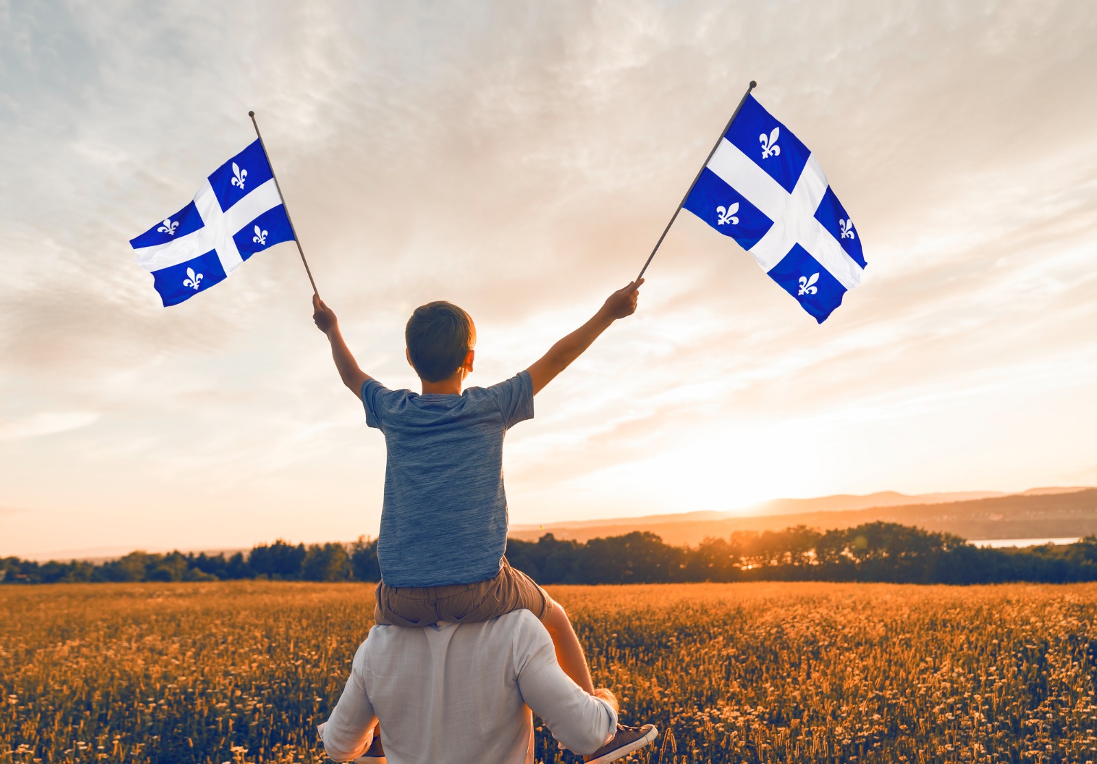 Père et enfant patriotes agitant des drapeaux québécois au coucher du soleil.