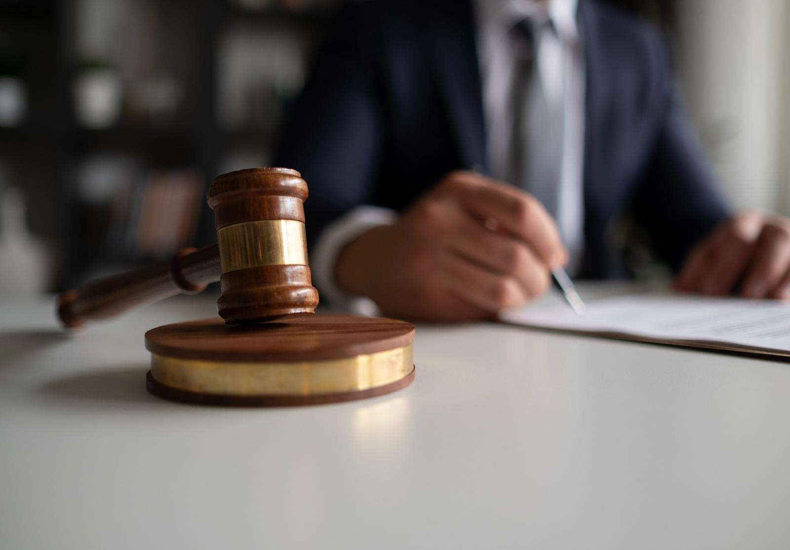 Marteau de juge en avant-plan, avec un homme en costume écrivant un document en arrière-plan.