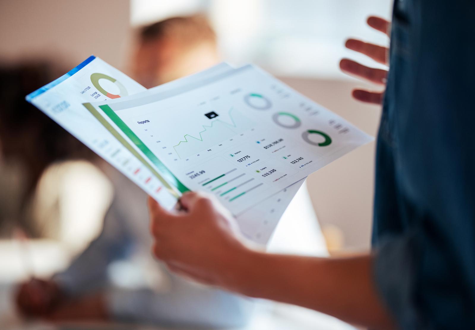 Personne tenant en main deux feuilles de rapports financiers imprimés, avec des graphiques (camemberts, courbes, barres).