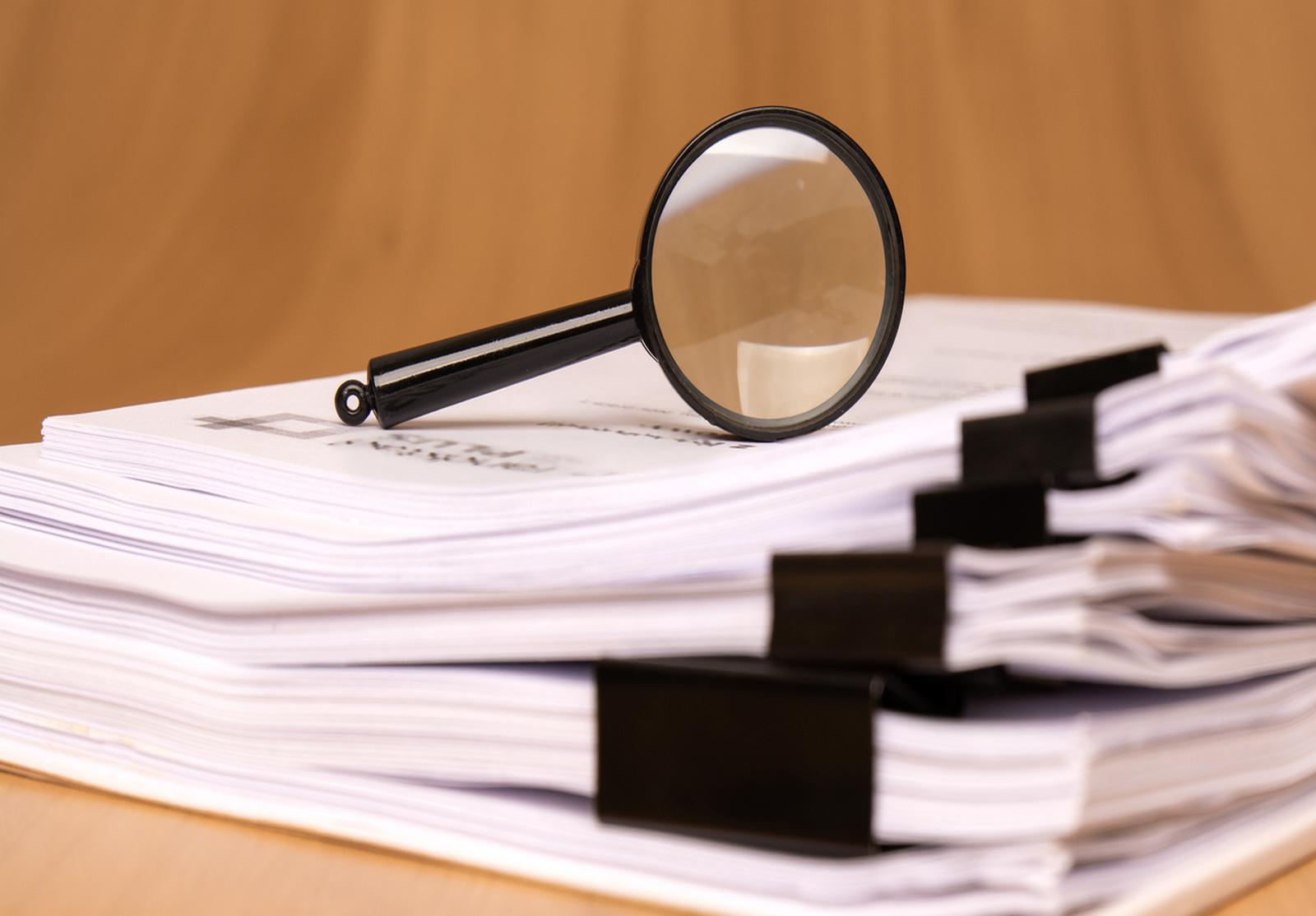 Gros plan sur une pile de documents reliés avec des pinces noires. Une loupe est posée au-dessus, suggérant l’idée d’analyse ou d’audit.