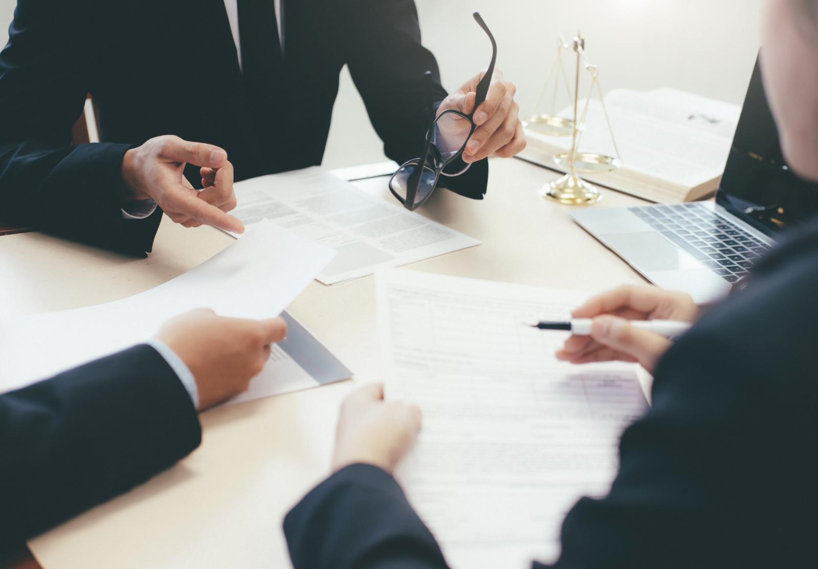 Réunion de travail entre personnes en costume, discutant et échangeant des documents juridiques, avec une balance dorée et un ordinateur portable sur la table.